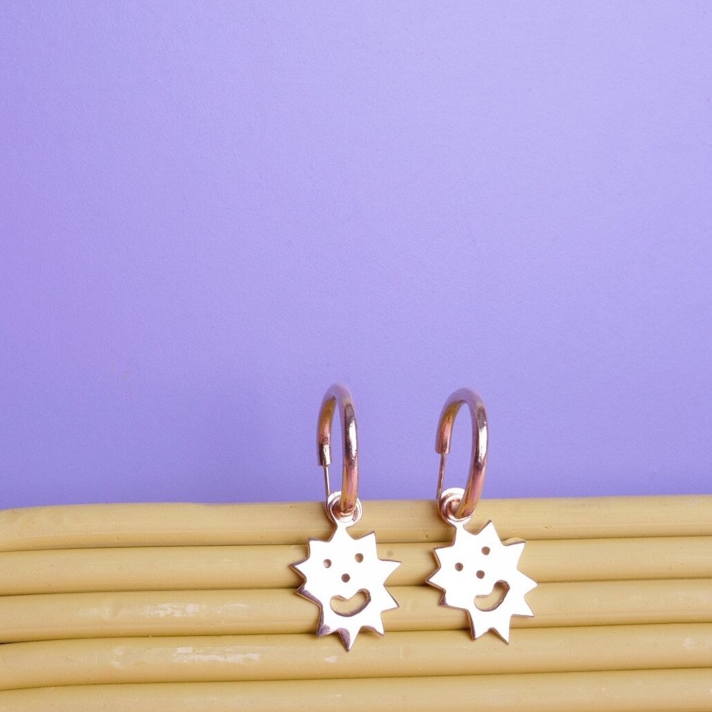  Small gold hoop earrings with sun shaped charms hanging on them. The charms have smiley faces cut out of them 