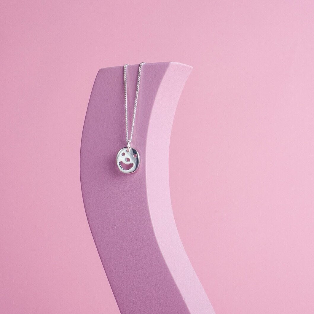  A silver pendant necklace with a charm hanging on it. The charm has a smiley face cut out of it.