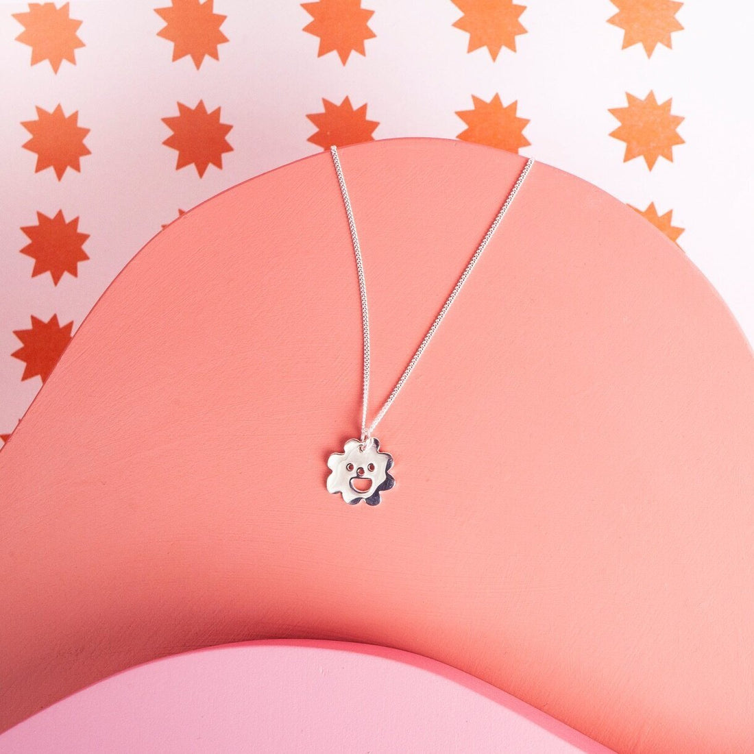  A silver pendant necklace with a flower shaped charm hanging on it. The charm has a smiley face cut out of it.