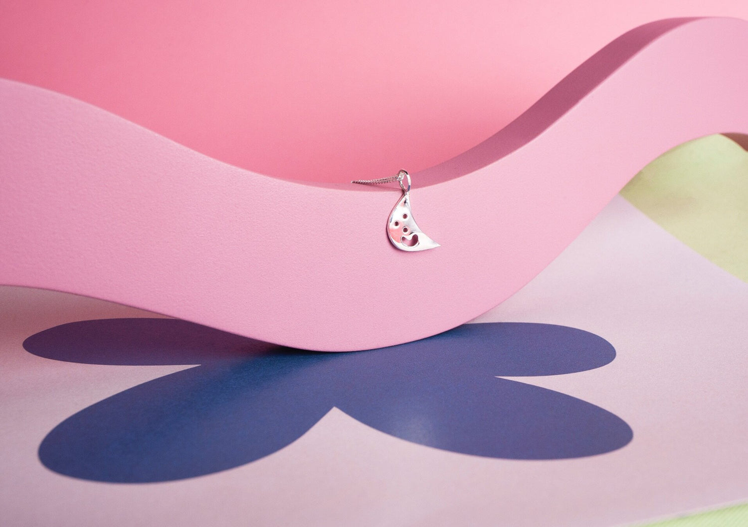A small silver moon shaped charm on a chain. The charm has a smiley face cut into it. The necklace is hanging over a pink abstract shape and there is a large purple flower shape printed n the table.